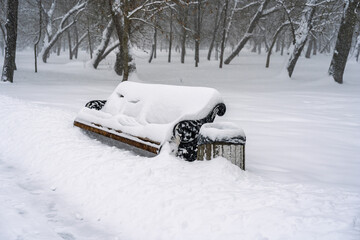 A wooden park bench and trash bin completely covered by heavy fresh white snow during a severe blizzard in the woodland