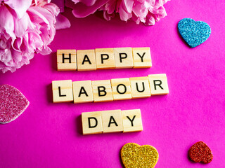 wooden blocks spelling happy labour day on pink background, pink peony and colourful hearts