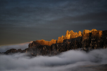 High-resolution landscape of jagged mountain peaks illuminated by golden hour light, rising above a dense layer of clouds under a dramatic dark sky.