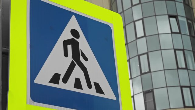 Close-up of a fluorescent yellow bordered pedestrian crossing sign with a blue triangular icon, set against the glass facade of a modern building, highlighting urban road safety.