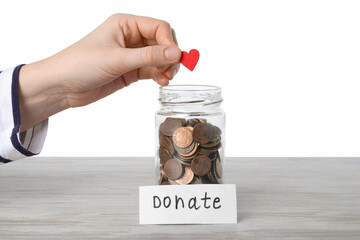 Woman putting red decorative heart into donation jar at wooden against white background, closeup