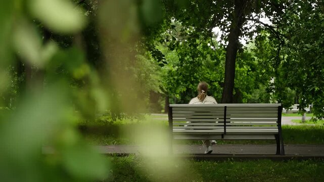 Solitary person sitting on a white park bench beneath lush green trees, viewed from behind through soft, blurred foliage, capturing a tranquil summer moment in a quiet leafy oasis