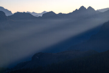 Dramatic mountain silhouettes during blue hour featuring a powerful sunbeam piercing through dense valley mist. Atmospheric landscape photography suitable for travel and wellness media.