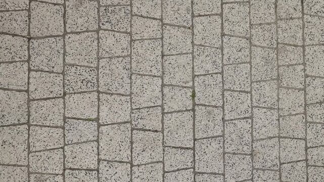 Overhead close-up of light gray cobblestone pavers arranged in staggered rows, with dark mortar joints and speckled stone texture creating a repeating geometric grid on a walkway.