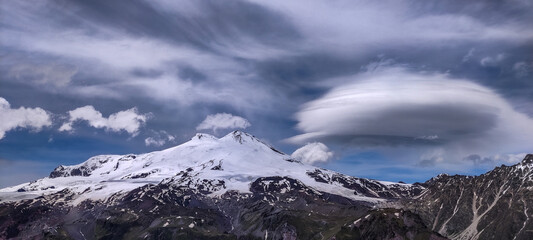 Panoramic landscape in the Caucasus Mountains with a view of Mount Elbrus in cloudy weather, lenticular clouds hanging in the sky above the mountains, glaciers and snow on the mountain