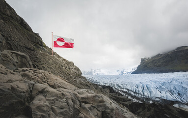Greenland national flag flies above a rocky Arctic glacier and mountains under a cloudy sky,...
