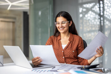 Young indian businesswoman in glasses reviewing financial documents with a laptop on her desk, smiling while working on business analytics and management