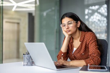 Young indian woman experiencing job stress and boredom, resting her head on her hand while working on a laptop at a modern office desk, feeling exhausted and procrastinating