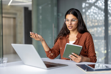 Young indian woman wearing glasses actively talking and gesturing during a remote video conference meeting, effectively collaborating from her modern office workspace while holding a notebook