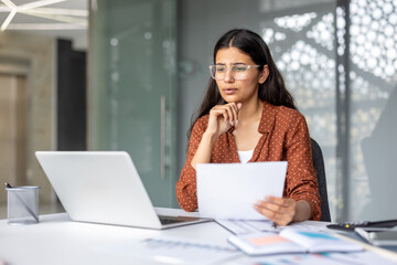 Young woman wearing glasses sitting at desk with laptop, holding document and concentrating while analyzing business data, budgets, or paperwork in a modern office setup