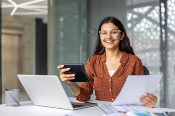 Young indian woman business professional smiling while sitting at a modern office desk, holding a...