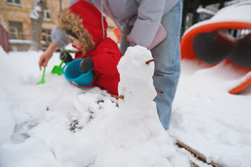 Child in bright red coat and parent enjoy winter by building a small snowman in a snowy playground. The child uses a bucket and shovel, creating joyful wintertime memories.