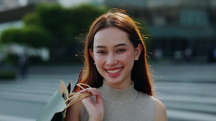 Portrait of joyful Asian woman with shopping bags on shoulder as stands on city square. Pretty Korean lady enjoys large sale on Black Friday in urban environment. Fashion trends