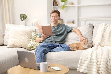 Smiling man reading book and stroking his cute cat on sofa at home © New Africa