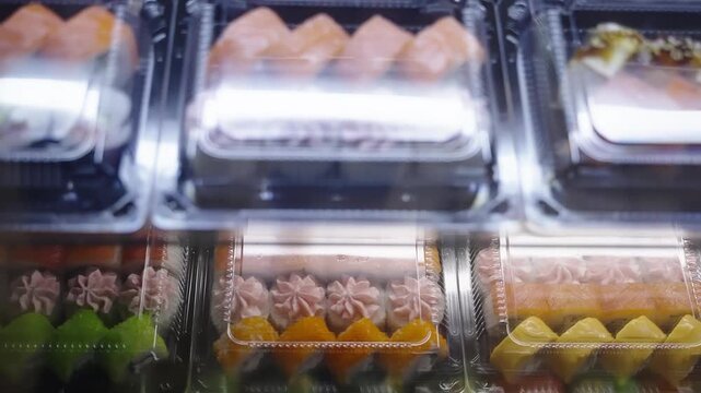 Assorted sushi in clear plastic clamshell containers displayed in a refrigerated case, featuring salmon nigiri and colorful rolls with sauces and garnishes, ready for takeaway