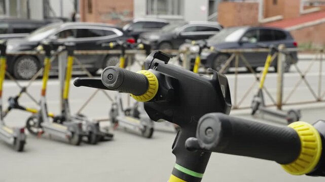 Close-up of an electric scooter handlebar with yellow grip accents, framed by a blurred row of parked e-scooters and cars along an urban street with railings and brick buildings.