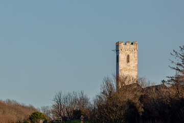 A medieval stone church tower with crenelated battlements rising above a winter treeline. This historic architectural landmark stands against a clear, pale blue sky in the evening light.