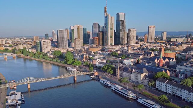 Aerial drone zoom-in shot from 100m over the Main River towards the Eiserner Steg bridge and the modern skyscraper skyline. Scenic urban landscape of the financial district in Frankfurt, Germany.