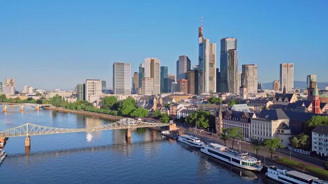 Aerial drone zoom-in shot from 100m over the Main River towards the Eiserner Steg bridge and the modern skyscraper skyline. Scenic urban landscape of the financial district in Frankfurt, Germany.