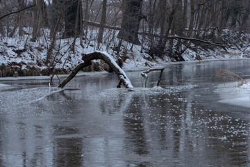 Mała rzeka zimą. Brzegi rzeki porastają krzaki i wysokie drzewa. Ziemię pokrywa warstwa śniegu, powierzchnia rzeki skuta jest lodem.