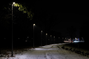  Ścieżka rowerowa obok gł&oacute;wnej drogi. Jest zima, nawierzchnia pokryta jest warstwą białego śniegu. Jest noc, ciemności rozświetla rząd lamp led. W ich świetle widać padający śnieg.
