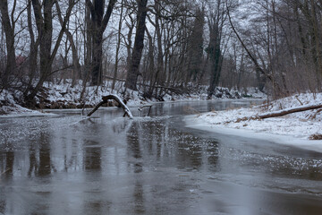 Mała rzeka zimą. Brzegi rzeki porastają krzaki i wysokie drzewa. Ziemię pokrywa warstwa śniegu, powierzchnia rzeki skuta jest lodem.