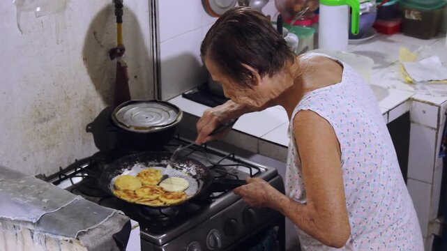 An elderly wrinkled Hispanic woman is grilling cornmeal arepas in a pan on a stove inside her home in Neiva, Huila, Colombia. Concept of healthy lifestyle