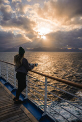Fototapeta premium Epic Sunset Cruise Ship Female Photographer Looks Over Balcony at Heavenly Sun Beams Through Clouds Over Ocean. Colorful Landscape Dreams Adventure