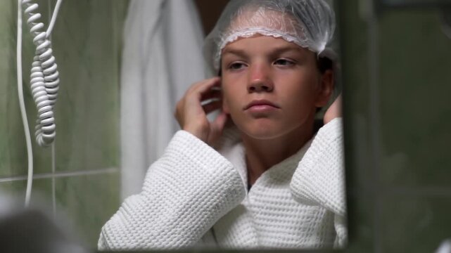 A girl looks at herself in a mirror in a hotel bathroom and adjusts the clear shower cap on her hair. The teenager is performing her morning or evening body care routine.