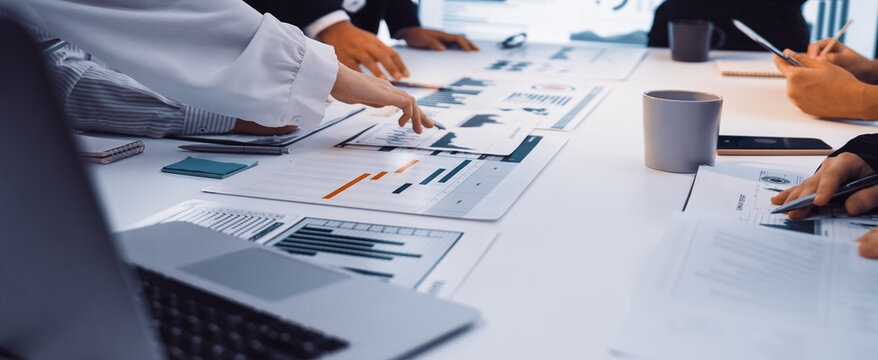 A business team engages in a collaborative meeting, analyzing financial reports and data charts laid out on a table in a modern office setting. SACTR - Powered by Adobe