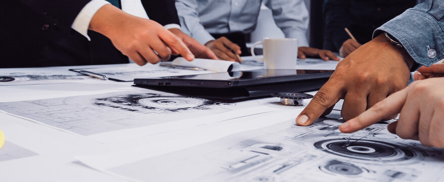 A diverse team collaborates on a technical project, discussing blueprints and using a tablet in a modern office to foster creativity and innovation. SACTR - Powered by Adobe