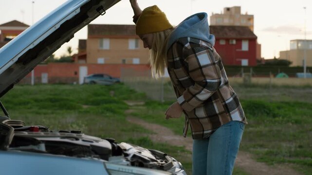 Stressed young woman stand on side of road with car hood open, inspecting engine and desperately trying to figure out what wrong with her broken down vehicle.Road problem, car trouble concept.