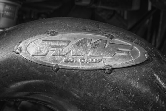Black and white close-up detail of a motorcycle exhaust pipe featuring a worn FMF metal badge. 
