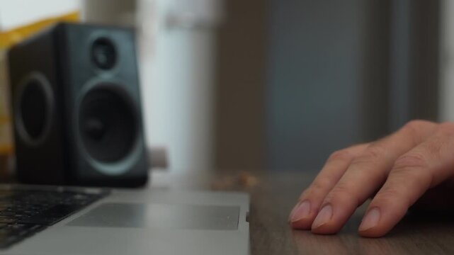 Close up of nervous hand tapping fingers on wooden desk next to laptop and speaker, showing impatience, boredom, or waiting for something important while listening to music