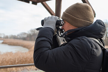 Caucasian male young adult birdwatcher in winter gear observing nature with binoculars