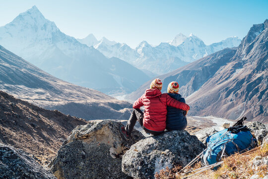 Couple resting on Everest Base Camp trekking route near Dughla 4620m. Backpackers left gear and trekking poles while enjoying valley view with Ama Dablam 6812m peak and Taboche 6495m.
