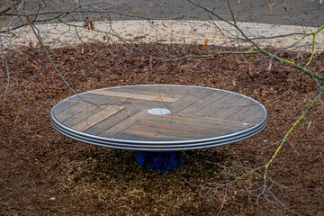 A Circular Wooden Platform With Metal Edging Sits Atop A Blue Base Surrounded By Brown Leaves And Bare Tree Branches