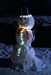 A Large Snowman Lit From Within By Glowing White And Orange Lights Wearing A Bucket Hat In The Night Snow