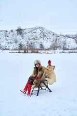 Fototapeta premium Stylish woman in fur coat, hat, and red boots sits on a chair in a snowy field with arm raised, creating a visually interesting contrast against the winter backdrop.