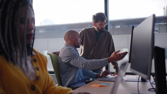 Diverse team gathers around a screen for a project meeting, exchanging ideas and planning next steps in a modern office environment with daylight and city skyline in the background.