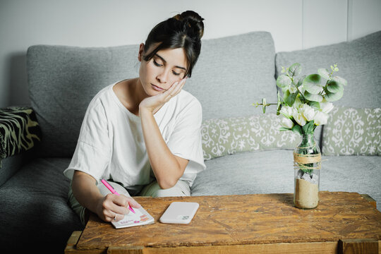Thoughtful young woman sitting on a sofa at home, writing notes on a notepad with a pen. Cozy minimalist living room interior with wooden coffee table, smartphone, and flowers in a glass vase