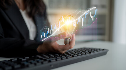 A professional holds a glowing light bulb while seated at a keyboard, symbolizing innovation in finance against a backdrop of digital data graphs and trends. Impute