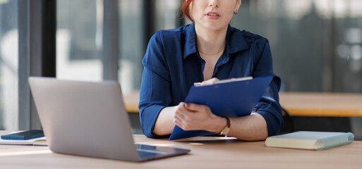 Middle aged serious woman working with laptop sitting at table make notes in copybook at office
