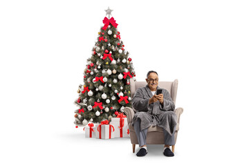 Mature man in a bathrobe sitting in an armchair and using a smartphone by the Christmas tree