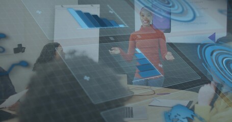 Presenting woman in red top and jeans gesturing at meeting table with augmented charts and laptops