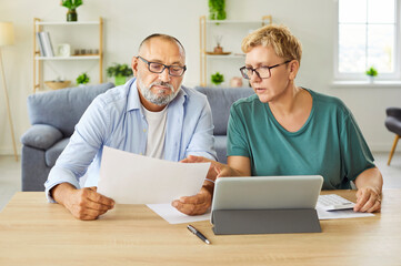 Senior couple sits at table with tablet and paperwork, discussing budget, expenses and retirement...