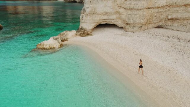 Woman running along curved cove beach past sea cave and emerald water on Greek island