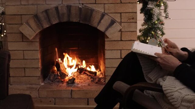 Woman writing in a notebook while sitting in an armchair by a burning fireplace. Cozy winter home interior with warm firelight and festive decor. Concept of slow living, planning, reflection, relaxati