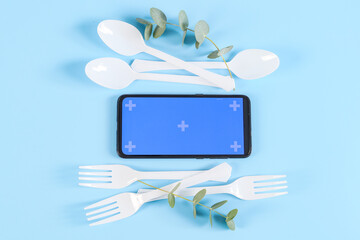 Mobile phone, plastic forks, knives and eucalyptus branches on a blue background.