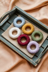 Fototapeta premium Rustic wooden tray with six knitted hair ties made of textured yarn in light blue, white, mustard yellow, deep red, green, and lavender, arranged on beige cloth over soft peach fabric.